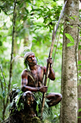 A man with painted skin and foliage adornments crouches in a forest, holding long spears and wearing a headdress with feathers, embodying the traditional attire of Papua New Guinea.