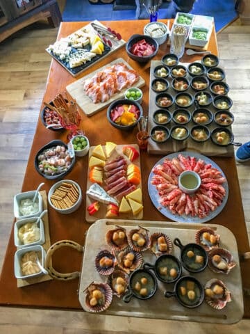 A wooden table is filled with an assortment of foods, including cheeses, meats, seafood, crackers, sauces, and fruit—all arranged in bowls and on platters reminiscent of a festive Argentine spread.