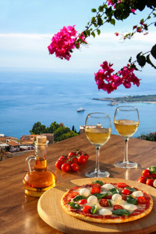 An outdoor table in Italy overlooks the ocean, adorned with two glasses of white wine, a bottle of olive oil, cherry tomatoes, and a pizza topped with tomatoes, mozzarella, and basil. Pink flowers bloom in the foreground.