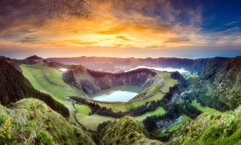 Panoramic view of a crater lake surrounded by green hills and a winding road at sunrise, under a partly cloudy sky.