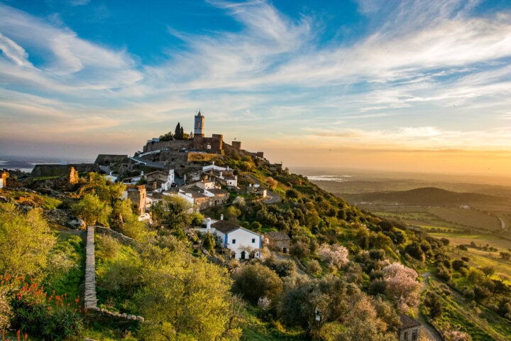 A hilltop village in Portugal with stone houses and a church near the top, surrounded by trees and greenery, under a partly cloudy sky during sunset. A winding path leads up the hill.