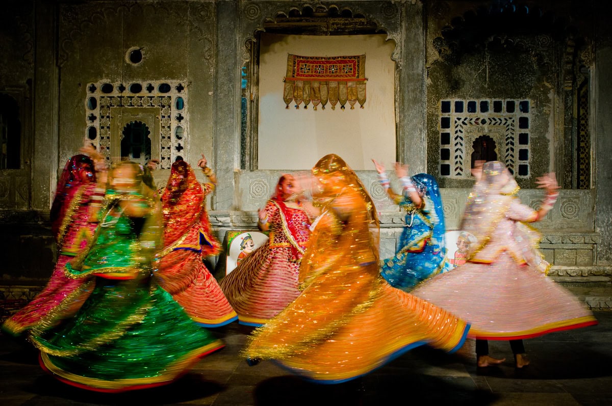 A group of women in colorful traditional attire perform a spinning dance in front of ornately decorated walls with arched windows—moments like these are not to miss in 2026.