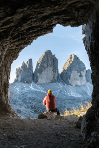 A person in a red jacket and yellow helmet sits at the entrance of a cave, facing a mountainous landscape with rocky peaks under a clear Italian sky.