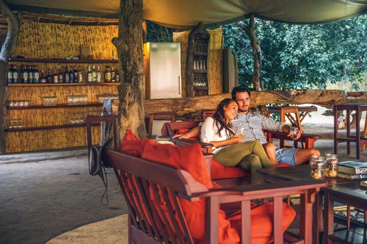A couple sits together on an outdoor lounge set under a canopy, with a rustic bar in the background, reminiscent of a cozy retreat in Zambia. They are both holding drinks and appear relaxed and content.