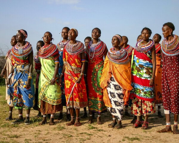 A group of individuals in traditional colorful attire and beadwork stands together outdoors, under a partly cloudy sky, as part of their memorable tour.