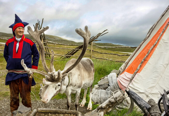 A person dressed in traditional clothing stands next to a reindeer near a tent-like structure in a grassy landscape with wooden fences in the background, capturing the essence of Finland's serene countryside.