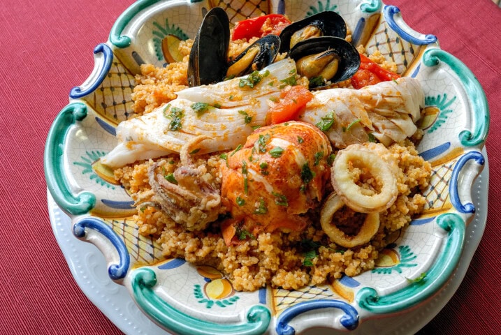 A colorful ceramic dish holds a seafood and couscous meal, featuring mussels, fish fillets, squid rings, and a lobster tail, garnished with herbs and tomato chunks on a red tablecloth reminiscent of Italy.