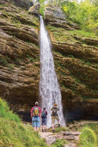 Three people stand at the base of a tall waterfall cascading down a rock face in Slovenia, surrounded by lush greenery.