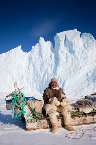 A person dressed in warm clothing sits on a wooden sled in a snowy landscape with large, jagged ice formations in the background, capturing the essence of travel through Greenland's stunning Arctic scenery.