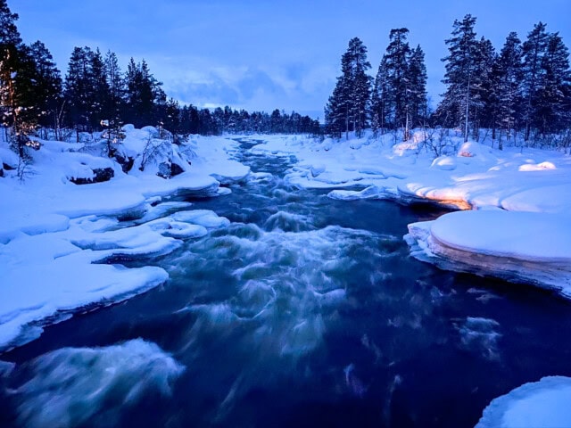 A river flows through a snowy landscape, surrounded by snow-covered trees under a twilight sky, capturing the serene beauty of Finland's culture and inviting tourism.