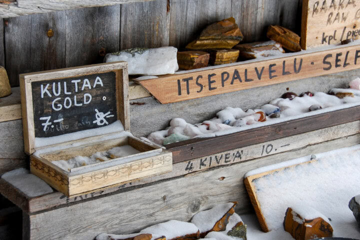 A wooden outdoor display, perfect for travel enthusiasts exploring Finland, offers various rocks and minerals for sale. Among the attractions, there's a small open box labeled "KULTAA GOLD" and other stones priced at €7 and €10, partially covered in snow.