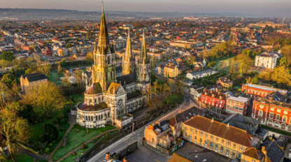 Aerial view of a large Gothic-style cathedral surrounded by trees and buildings in a cityscape at sunrise, highlighting one of the must-see Cork attractions for those exploring Cork tourism.