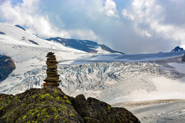 A cairn of stacked stones stands on a mossy rock with a backdrop of Switzerland's snowy glacier and mountains under a partly cloudy sky.