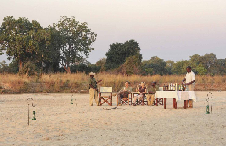 Four people sit in folding chairs around a small table with drinks, attended by two servers, in a sandy outdoor area in Zambia with trees and grass in the background. Lanterns are placed on the ground.