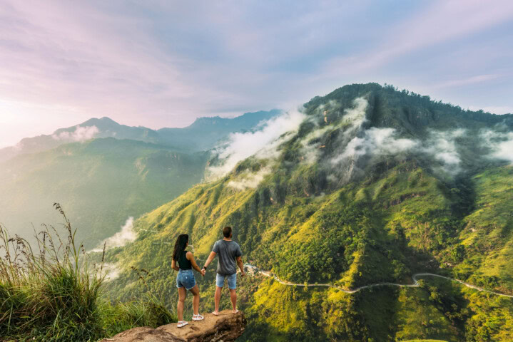 Two people stand hand in hand on a cliff edge, overlooking a misty mountainous landscape in Sri Lanka, with green vegetation and winding roads—a perfect snapshot of travel and tourism. The sky is partly cloudy.