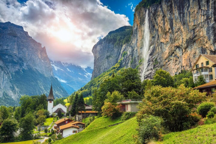 A village with a church and houses sits in a mountainous valley of the Swiss Alps. A waterfall cascades down a cliff beside greenery, under a partly cloudy sky with sunlight breaking through. This idyllic location is an ideal travel destination in Switzerland.