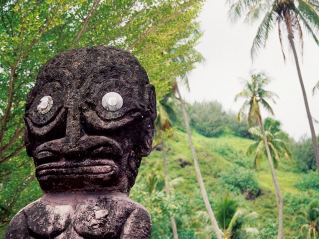 A stone statue with large eyes and pronounced facial features stands in front of lush green tropical foliage and palm trees, evoking the mystique of French Polynesia.