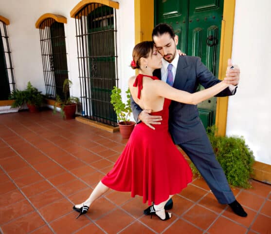 A man and woman perform a tango dance indoors on a terracotta-tiled floor, with the woman in a red dress and the man in a gray suit, capturing the passionate spirit of Argentina's rich culture—a perfect moment for any travel enthusiast exploring this vibrant destination.