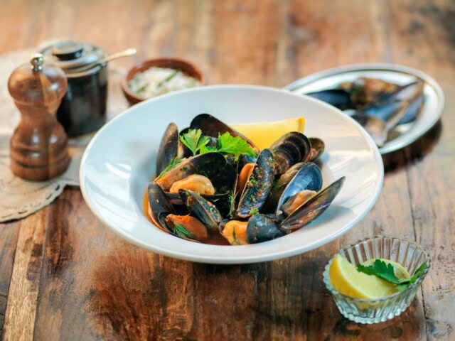 A plate of cooked Tasmanian mussels garnished with herbs and a lemon wedge, served with additional lemon slices and a pepper grinder on a wooden table.