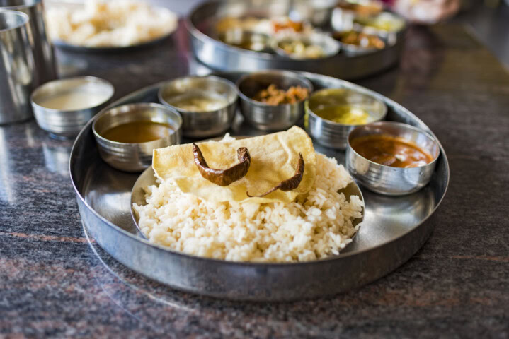 A stainless steel plate holds white rice, a piece of papadum, and small bowls of various Sri Lankan and Indian curries and sauces.
