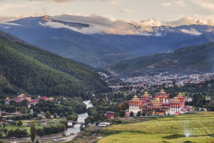 A scenic view of a Bhutan valley with a river and vibrant temples surrounded by green forests and fields, set against a backdrop of cloud-topped mountains—an enchanting destination for travel enthusiasts.