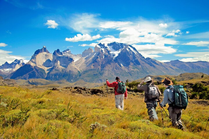 Three hikers with backpacks and trekking poles walk on a grassy trail towards the snow-capped mountains of Chile under a blue sky with scattered clouds.