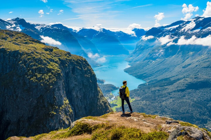 A person stands on a cliff edge overlooking a vast Norwegian mountainous landscape with a serene lake below, under a clear blue sky.