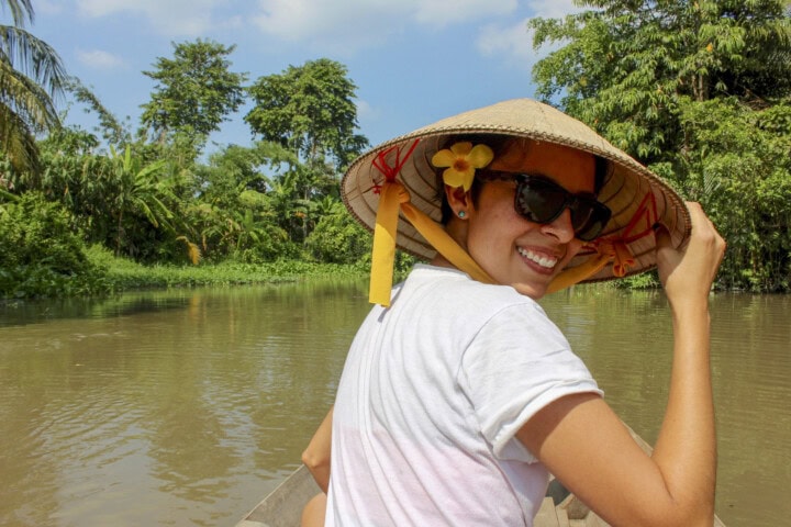 Person in a white shirt and sunglasses wearing a conical hat, smiling while sitting in a boat on a river surrounded by lush greenery, capturing the essence of Vietnam travel.