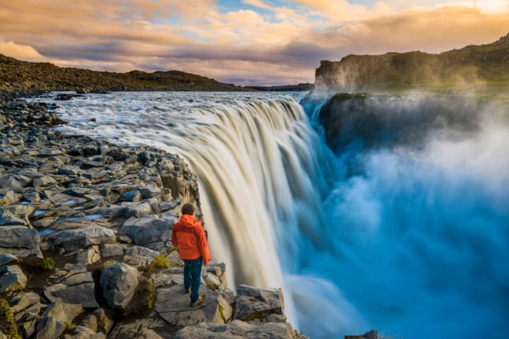 A person in a red jacket stands on a rocky cliff side, overlooking a large waterfall cascading into a gorge under a partly cloudy sky, capturing the spirit of travel and tourism in Iceland.