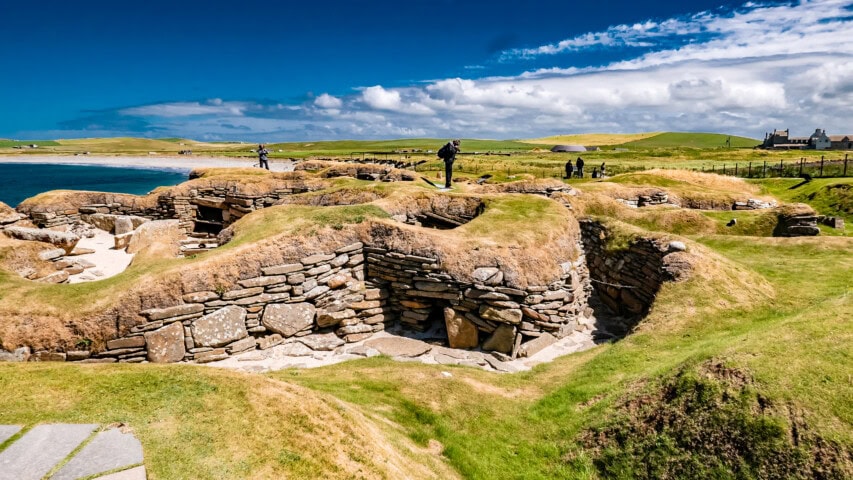 A group of visitors explores the ancient stone structures and grassy mounds at Skara Brae, a Neolithic village on the Orkney Islands in Scotland, under a sunny, blue sky.