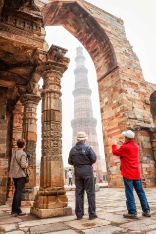 Three travelers stand under ancient stone arches with ornate carvings, as they view and photograph a tall, historic sandstone tower in the background.