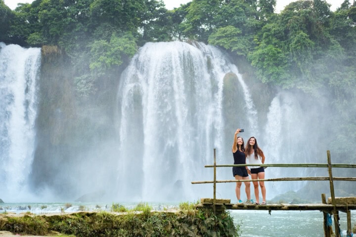 Two people stand on a wooden platform, posing for a selfie in front of a large waterfall surrounded by lush greenery—a perfect travel moment epitomizing the beauty of Vietnam’s tourism.