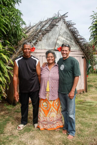 Three people stand together smiling in front of a small, rustic wooden building with a thatched roof, reminiscent of the charming architecture found in Tonga.