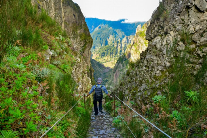 A person with a backpack hikes a narrow, rocky mountain path surrounded by lush greenery and steep cliffs, with a valley and distant mountains in the background—a perfect setting for travel enthusiasts exploring the scenic landscapes of Portugal.