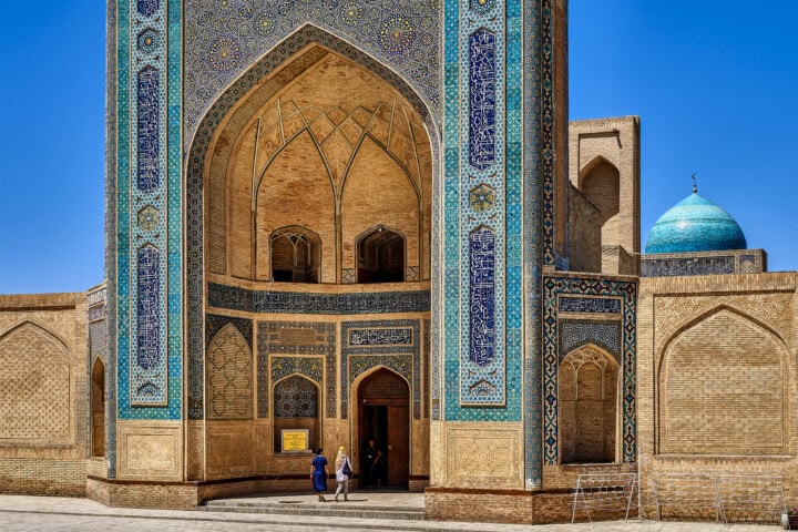 Two people stand at the entrance of a large, ornate mosque in Uzbekistan, its intricate blue tile work and brick walls showcasing stunning craftsmanship.