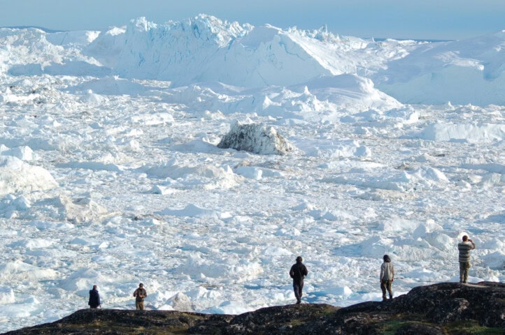 Five people stand on a rocky hill in Greenland, overlooking a vast, icy landscape filled with snow and ice formations under a clear sky.