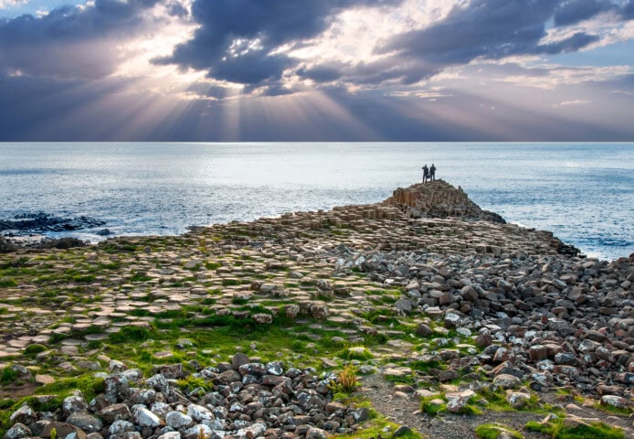 Two people stand on the rocky Giant's Causeway formation in Ireland, facing the ocean, with sunlight breaking through clouds in the background—a perfect moment that captures the essence of travel and tourism.