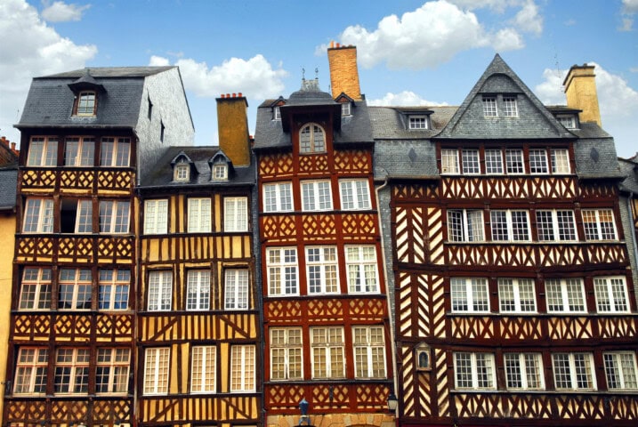 A row of traditional half-timbered houses with steep roofs and multiple windows under a bright blue sky with scattered clouds, reminiscent of picturesque French villages.