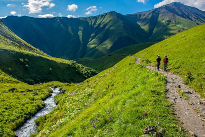 Two people hike along a narrow trail beside a small stream in Georgia's lush, green mountainous landscape under a partly cloudy sky.