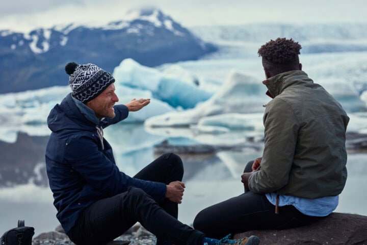 Two men sit on rocks by a glacier, with one gesturing toward the icy landscape. Snow-covered mountains and large ice formations are visible in the background.