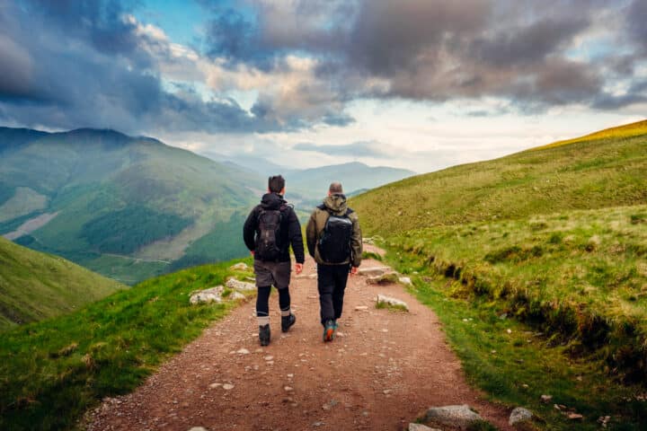 Two people with backpacks walk on a dirt trail through the green, hilly landscape of Scotland under a cloudy sky.