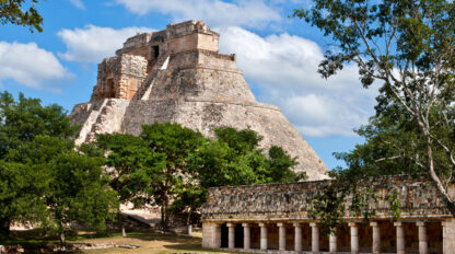 Ancient stone pyramid with steep steps surrounded by trees and smaller stone structures under a clear blue sky, unlocking the secrets of the Maya civilization.