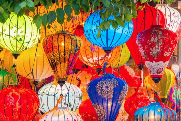 A collection of colorful lanterns, reminiscent of vibrant Vietnam markets, hangs from tree branches. The red, blue, yellow, and green lanterns showcase various patterns and designs.