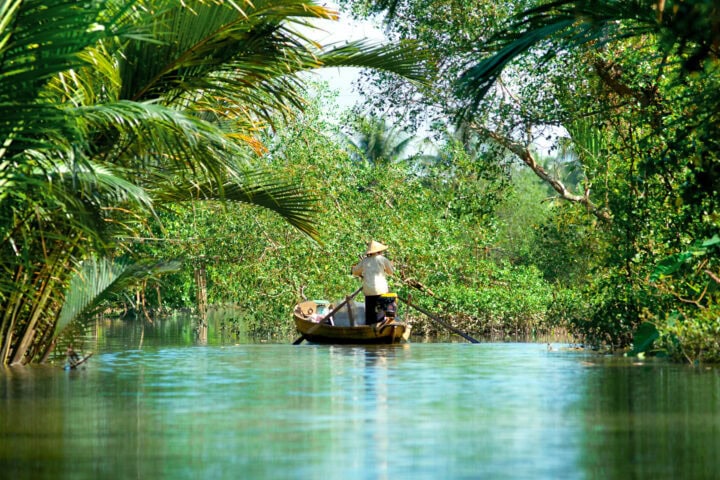 A person wearing a conical hat rows a wooden boat through a narrow, lush, green waterway surrounded by dense tropical vegetation.