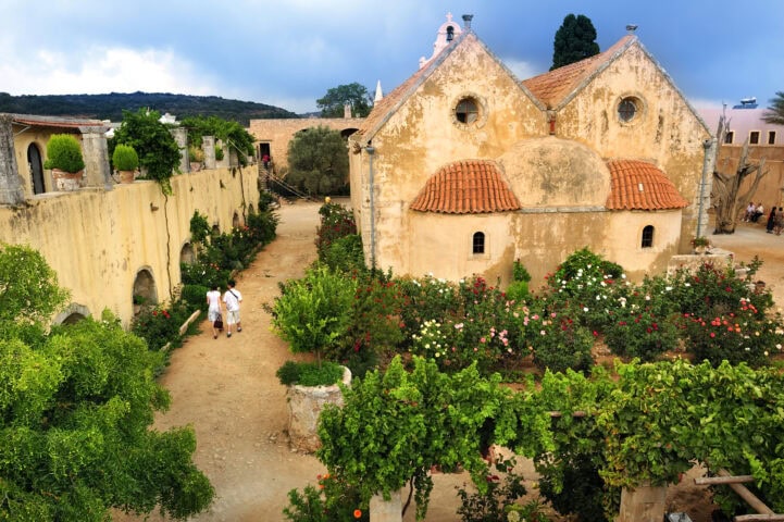 Two people walk through a garden with flowering bushes and trees beside an old, rustic building with red-tiled roofs.