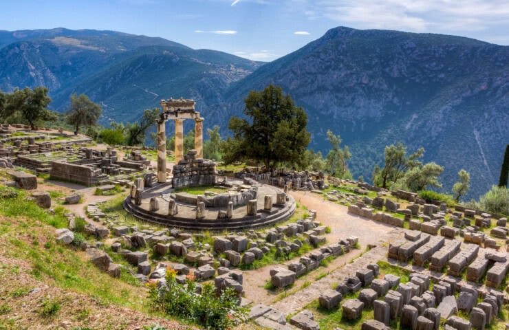 Ruins of the Tholos of Delphi with Greece's mountainous landscape in the background. Stone columns and circular structure are visible among scattered stone remnants and lush greenery.