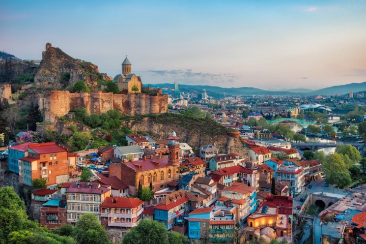 Panoramic view of Tbilisi, Georgia featuring historic buildings with colorful rooftops, cobblestone streets, and the ancient Narikala Fortress situated on a hill in the background. The charm of Georgia's capital city is truly undeniable.