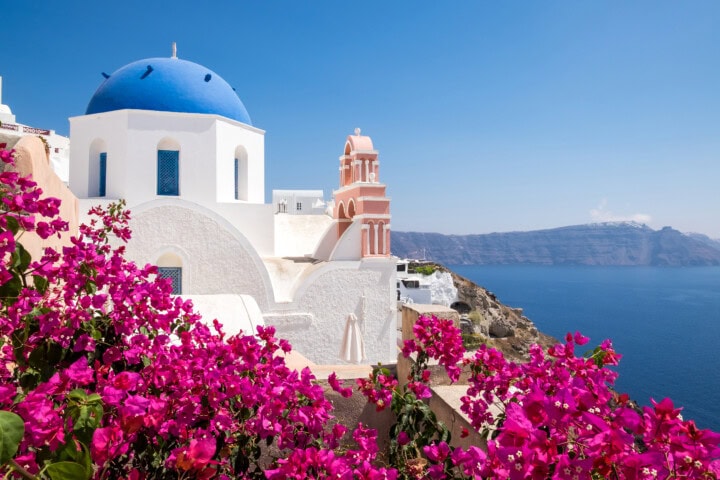 White buildings with a blue dome, a bell tower, and vibrant pink flowers in the foreground overlook a blue sea with distant islands under a clear Greece sky.