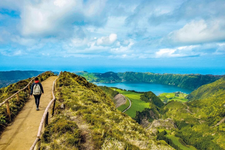 A person walks along a narrow dirt path atop a grassy hill in Portugal, with a vibrant blue lake and green landscape in the background under a partly cloudy sky.