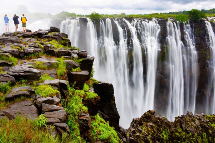 Three travelers stand on rocky terrain near the edge of a large, cascading waterfall surrounded by lush greenery, pausing to admire the view during their hike.